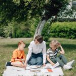 A woman teaches two kids outdoors on a picnic blanket under a tree.