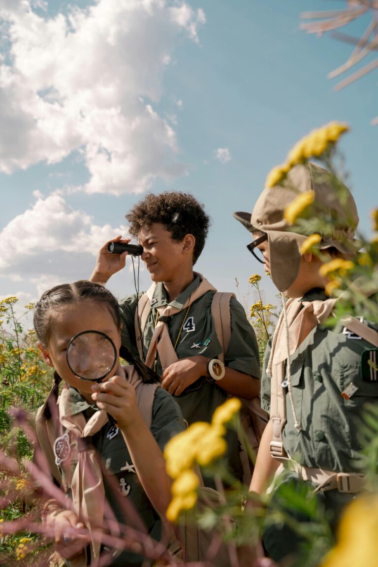 Three children in scout uniforms exploring nature with binoculars and magnifying glass on a sunny day.daycare centre nature based learning in early childhood wildlife facts for children